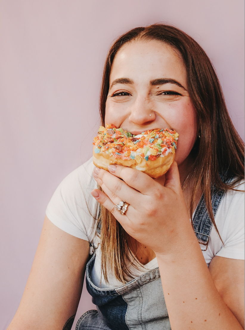 Photo of Alexandra laughing and biting into a donut covered in rainbow Fruity Pebbles. She is in overalls with a light pink background. This represents Alexandra's rebelling against diet culture.